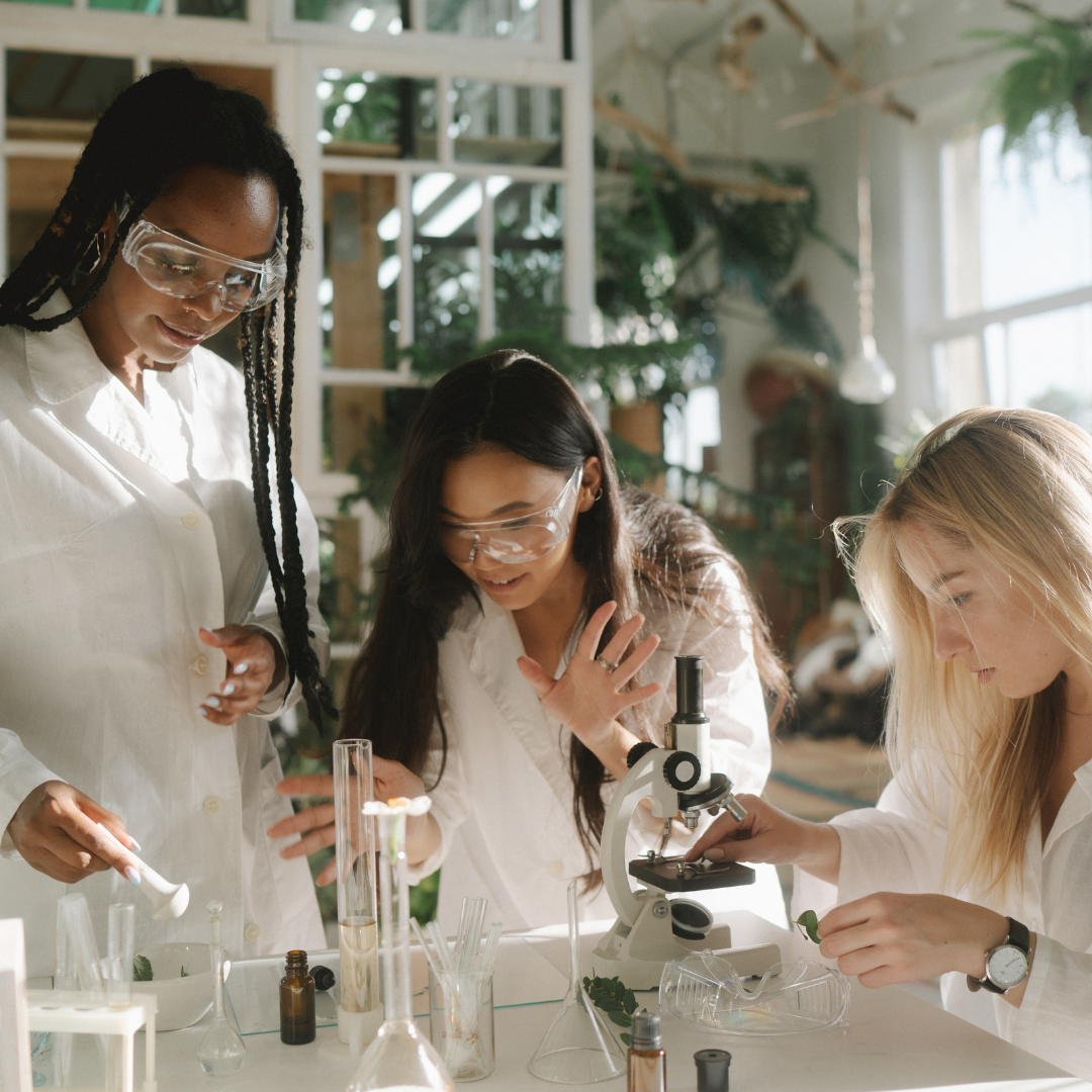 stock photo of a black woman with braids, an asian woman and a blonde woman wearing clear safety goggles and white lab coats huddled around various lab equipment 